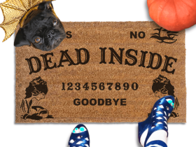 a Halloween welcome mat with a pug in golden dragon wings costume, the word "DEAD INSIDE" on it. The mat has a Ouija board design and the numbers "1234656789 GOODBYE" printed on it. The scene includes a person's feet wearing black pointy toed witch boots, an orange pupkin, and the text "DAMN GOOD DOORMATS EST. 2009" is visible.