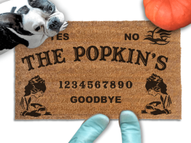 a french bulldog standing on a welcome mat with a custom last name on it. The mat has a Ouija board design and the numbers "1234656789" printed on it. The scene includes a person's feet wearing blue boots, a Halloween pumpkin, and the text "DAMN GOOD DOORMATS EST. 2009" is visible.