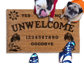 a welcome mat with a french bulldog and a fawn pug in bright hoodies, the word "UN-WELCOME" on it. The mat has a Ouija board design and the numbers "1234656789" printed on it. The scene includes a person's feet wearing white shoes, a vintage Halloween skull and cat light, and the text "DAMN GOOD DOORMATS EST. 2009" is visible.