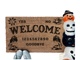 a welcome mat with the word "WELCOME" on it. The mat has a Ouija board design and the numbers "1234656789" printed on it. The scene includes a person's feet wearing white shoes, a vintage Halloween skull and cat light, and the text "DAMN GOOD DOORMATS EST. 2009" is visible.