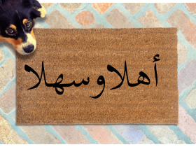 a dog peeking over a brown doormat with Arabic script on it reading welcome