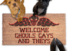 a humorous LGBTQ Halloween door mat with the text "Welcome Ghouls, Gays and Theys" and terrier and a black pug on top of it. There is also a pumpkin and a person's feet visible in the image.