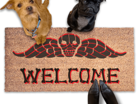 a dog and a small black pug and a terrier sitting on a welcome mat with a Beetlejuice skull design. The mat has the word "WELCOME" written on it in bones lettering