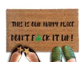 Coir doormat reading "This is our happy place, don't fuck it up," with a green cannabis leaf replacing the U in FUCK. Lifestyle photo shows two pairs of feet standing at the front edge of the mat.