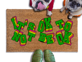 A pug and french bulldog in hoodies standing on a doormat with the text "IT'S OK TO BE OK" written on it, along with a person's feet wearing green shoes.