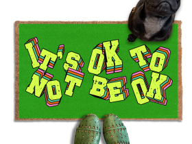 A pair of green shoes and a black pug dog are featured on a green doormat with the text "It's OK to BE OK" written on it.