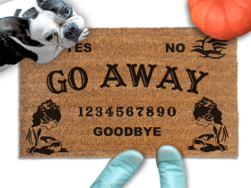 a Halloween welcome mat with a french bulldog, the word "GO AWAY" on it. The mat has a Ouija board design and the numbers "1234656789" printed on it. The scene includes a person's feet wearing Smurf blue boots and the text "DAMN GOOD DOORMATS EST. 2009" is visible.