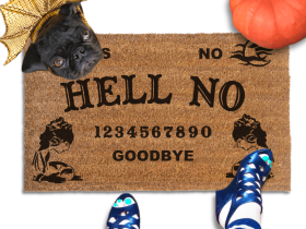 a Halloween welcome mat with a pug in golden dragon wings costume, the word "HELL NO" on it. The mat has a Ouija board design and the numbers "1234656789 GOODBYE" printed on it. The scene includes a person's feet wearing black pointy toed witch boots, an orange pupkin, and the text "DAMN GOOD DOORMATS EST. 2009" is visible.