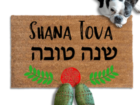 coir doormat reading Shana Tova in english and hebrew and a decorative pomegranate and wreath border.with a person in green slipons and a french bulldog