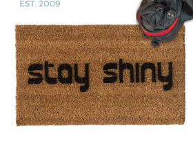 Natural coir doormat with black lettering reading Stay Shiny, Firefly, pictured with a black pug in a red harness looking up. Nerdy sci-fi welcome mat.