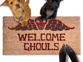 a terrier and a black pug dog sitting on a door mat with the text "WELCOME GHOULS" and a spooky Beetlejuice tombstone on it.