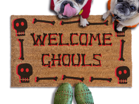 A french bulldog and a fawn pug in hoodies peeking over a welcome mat with the text "WELCOME GHOULS" and Halloween skull-themed decorations.